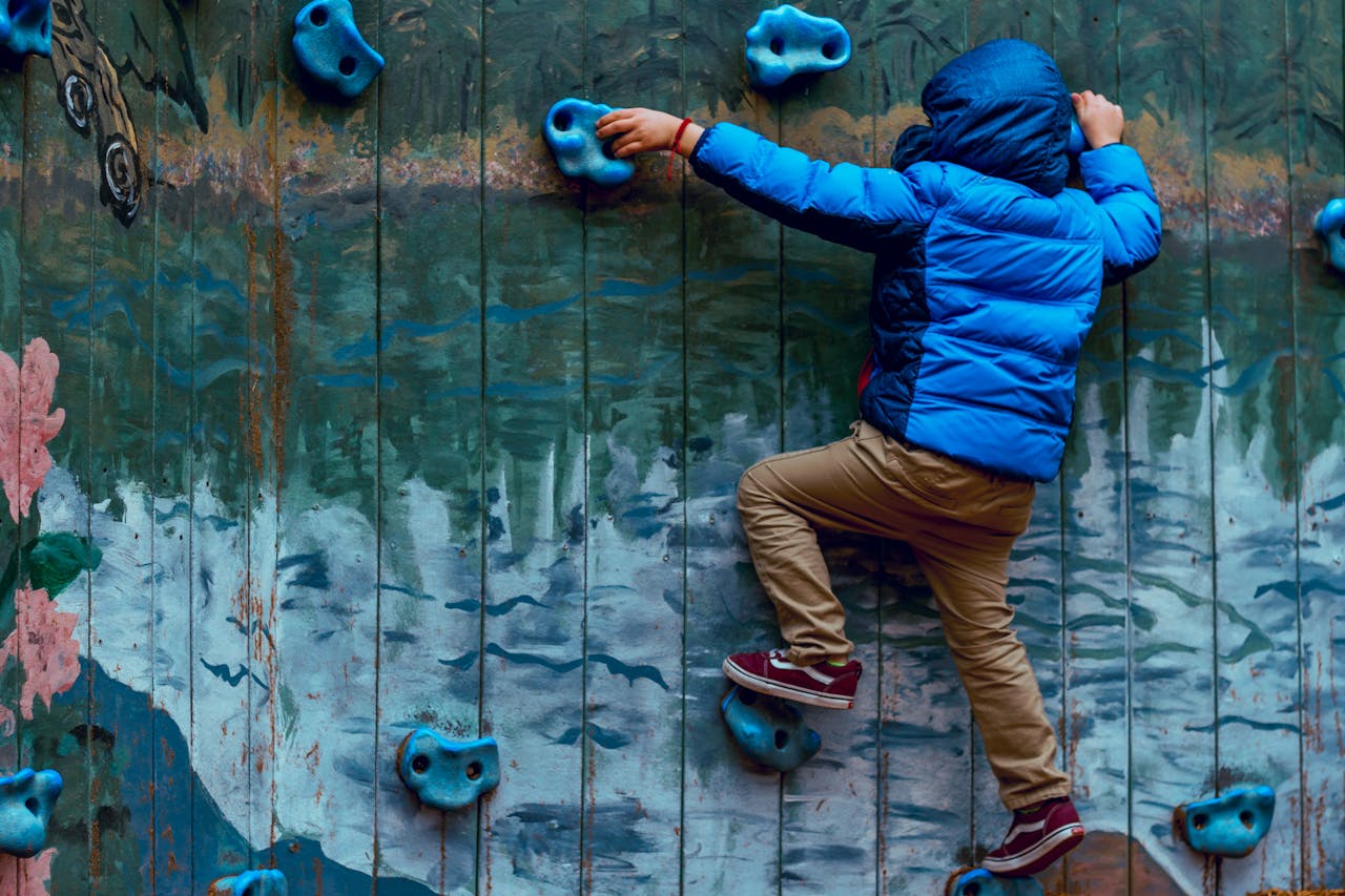 our-story Young boy in blue jacket climbing a painted rock wall outdoors, showcasing adventure and play.
