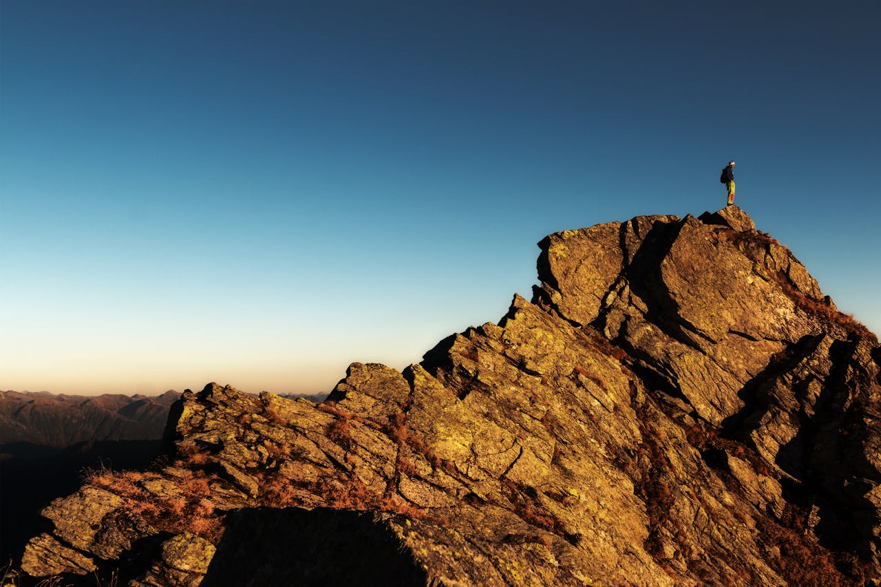 A lone hiker stands triumphantly atop a rocky mountain peak at sunrise, embracing nature and the view.